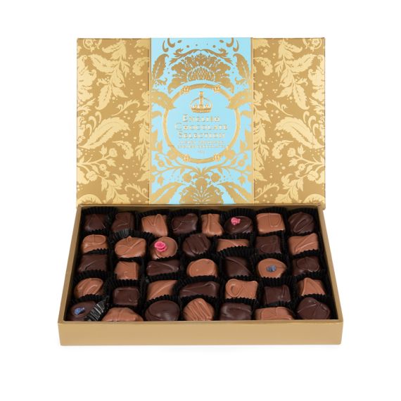 A side angle of the box of English Truffles with gold embossed Acanthus packaging and a light blue band with "English Truffles: Luxury Handmade English Chocolates." The tray of mixed chocolates are positioned at the bottom, with violet and English rose cr A side angle of the box of English Truffles with gold embossed Acanthus packaging and a light blue band with "English Truffles: Luxury Handmade English Chocolates." The tray of mixed chocolates are positioned at the bottom, with violet and English rose cr